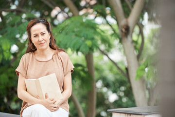 Naklejka premium Pensive mature woman sitting in park with opened book thinking after finishing reading
