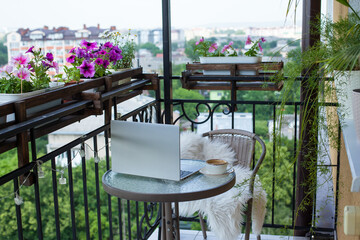 Laptop and cup of coffee on the table on the cozy balcony with flowers at home with city view. Remote work, doing business on vacation. Fast Internet, communication on weekends. Always in touch. 