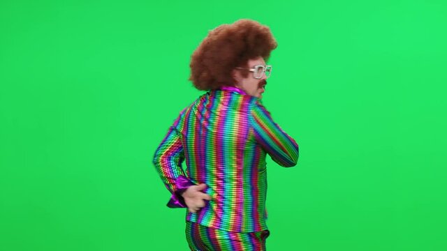 A retro dude in a funny wig and multicolored suit is dancing a funny spinning dance on a green background, a festive mood, a retro disco man is having fun at a new year's party.