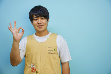青背景の前で手で丸を作るエプロンを着たアジア人男性	

