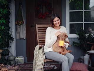 Young asian woman resting outdoors with cup of coffee on porch of country house, chilling outside with poodle dog . autumn lifestyle, leisure free time concept. Copy space