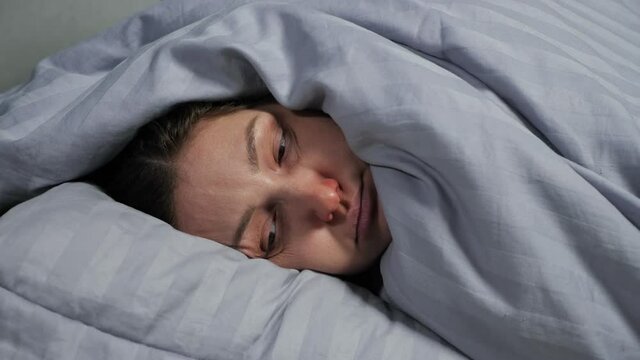 Young brunette woman with runny nose can not fall asleep because of severe cough and lies covered from head to toe with warm blanket closeup.