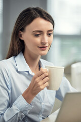 Attractive brunette Caucasian businesswoman in shirt using laptop and drinking coffee from mug in office