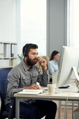 Serious young male helpdesk operator with beard adjusting headset microphone and answering call of client in office