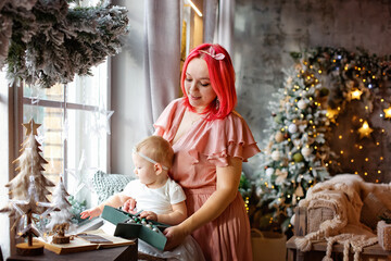 A mother with pink hair and a daughter are sitting on a windowsill in a beautifully decorated New Year and Christmas room against the background of lights of garlands.