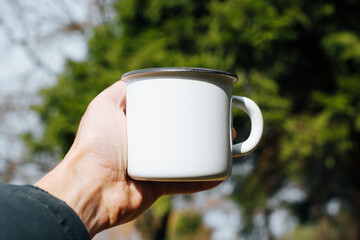 Concept hike, camping, trip. Hand showing empty white enamel mug with blank space for text or...