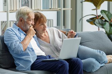 Caucasian old senior elderly grandparents couple sitting cuddling hugging together on sofa in living room at home using credit card and laptop computer browsing internet shopping from online store