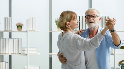 Obraz premium Caucasian old senior elderly happy grandparents lover married couple standing smiling listening to slow waltz music dancing together in living room at home. Gray hair bearded husband hold wife hand
