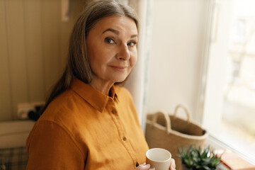 Close up shot of beautiful senior lady with long gray hair standing by window with cup of morning...