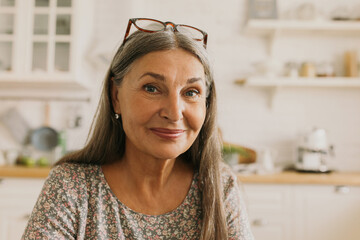 Pretty grey-haired elderly woman keeping glasses on forehead posing in front of camera with...