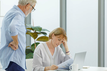 Caucasian old senior elderly stress depress worry female housewife reading financial saving insurance data problem have headache while grey bearded husband standing supportive in blurred foreground