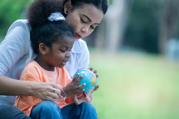 Happy Mother teaches daughter about our earth at park, dark skinned girl holds globe model in hands