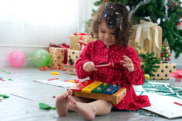 Cute little girl play wooden toy xylophone on floor, Christmas tree and gift boxes background,