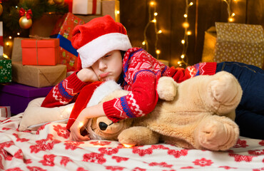 Child girl posing and dreaming in new year or christmas decoration. Holiday lights and gifts, Christmas tree decorated with toys. She's wearing a red sweater and a Santa helper hat