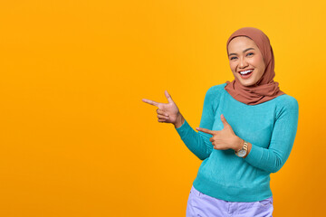 Portrait of smiling young Asian woman pointing finger at copy space on yellow background