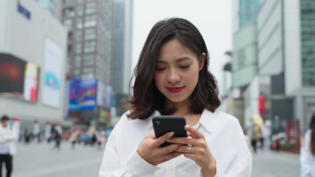 front view Charming young asian business woman walking in the city street looking at mobile phone in hand happy Chinese young lady using smartphone in urban background 4k slow motion lifestyle footage