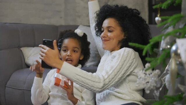 Portrait of African American mom and her little daughter communicate by video call using smartphone. Woman and girl sitting near sofa and decorated Christmas tree. Close up. Slow motion.