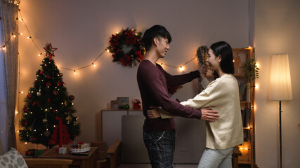young beautiful asian couple smiling happy and confident standing and dancing around christmas tree at home at night. sweet girl and boyfriend holding hands and doing social dance in xmas eve