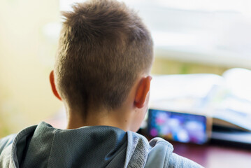 Distance learning online education. A schoolboy 10s boy studies at home and does school homework.Child sitting at table with smart phone,does remote homework.Selective soft focus blurred background.