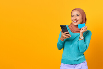 Naklejka premium Smiling young Asian woman holding credit card and smartphone while looking up on yellow background