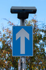 A traffic sign - while arrow pointing upwards on a blue background, with a light above, mounted on a pole in a one-way street