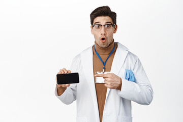 Image of surprised doctor pointing at mobile phone screen horizontal, showing application on smartphone, standing over studio background