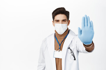 Healthcare worker in face mask from covid 19, wearing rubber gloves, showing stop no gesture, taboo and prohibition sign, standing over white background