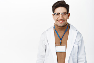 Fototapeta premium Medical workers and healthcare. Smiling male doctor in glasses and name tag, looking happy and confident at camera, standing over white background
