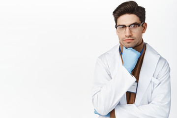 Handsome male doctor in medical robe and gloves, looking thoughtful, thinking of smth, standing against white background