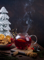 Winter hot tea with dried lemons, berries and spices in glass teapot with steam on wooden desk,  dark background. Italian biscuits cantucci.