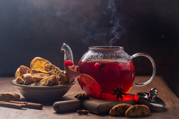 Autumn or winter hot tea with lemons, berries and spices in glass teapot with steam on wooden desk, dark background.Italian biscuits cantucci.