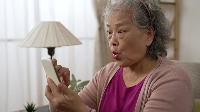 surprised asian elderly woman is exclaiming over the shocking online news on the phone while she is scrolling on the screen to check details in the living room