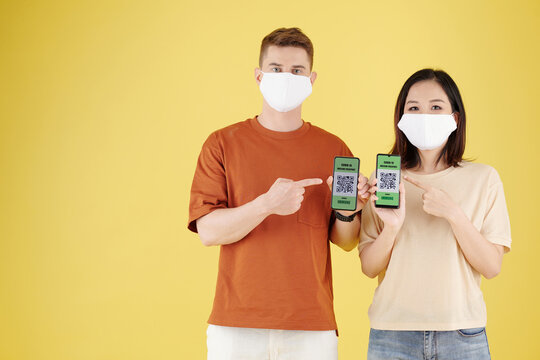 Diverse young couple pointing at QR codes on smartphones after getting vaccines against covid-19, isolated on yellow