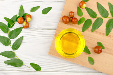 Jojoba oil in a bowl, a sprig of jojoba and fruits on a wooden table, top view