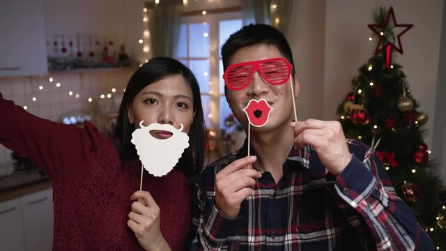 happy asian husband and wife looking at camera while taking photos of themselves holding funny Christmas masks on stick in a festive home interior at night.