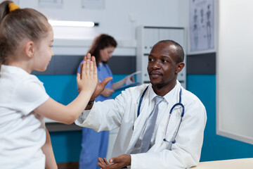 African american pediatrician doctor giving high five to young children during medical appointment in hospital office. Therapist man explaining illness symptoms discussing healthcare treatment