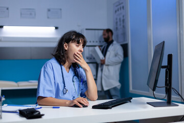 Exhausted nurse using computer on desk while falling asleep, working late. Medical assistant...