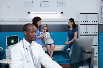Fototapeta premium African american pediatrician doctor typing diagnostic expertise on computer during medical examination in hospital office. In background physician nurse writing patient disease symptoms on clipboard