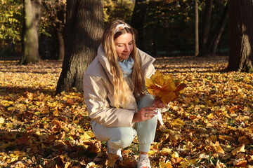 a young slender girl in a beige down jacket collects autumn fallen yellow leaves in the park on an...