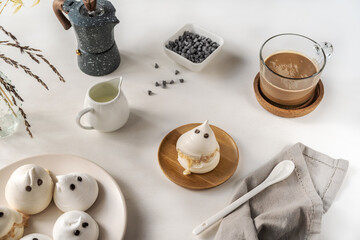 Meringues in a form of ghosts with cup of coffee, milk jar, chocolate drops and coffe pot on the light background