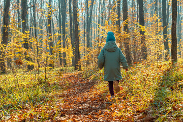 A girl child in a jacket and cap is running through the autumn forest. View from the back. In the background is green grass, yellow leaves and tree trunks. The concept of a happy childhood.
