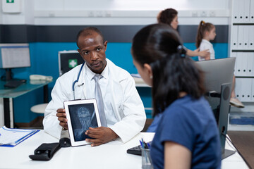 African american pediatrician doctor holding tablet computer explaining radiography expertise to mother during clinical consultation in hospital office. Bones xray on screen. Health care service