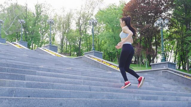 Slow motion sporty woman jogging up on stairs in park on sunny morning. Tracking shot of female jogger from behind exercising outside. Concept of fitness