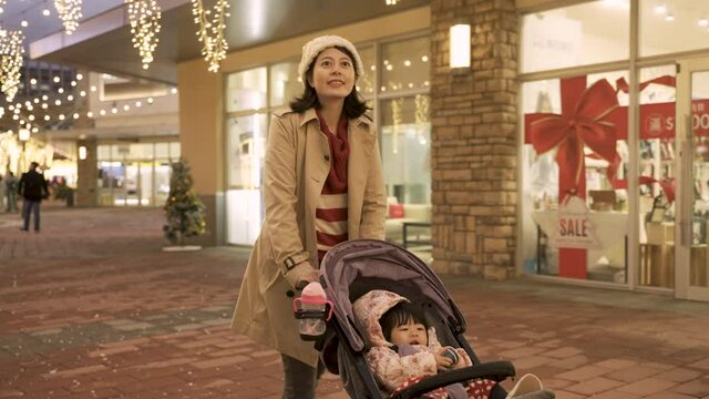 slow motion cheerful asian mother feeling joy while walking in the urban area shopping for Christmas gifts on a snowy evening with her baby girl in the stroller