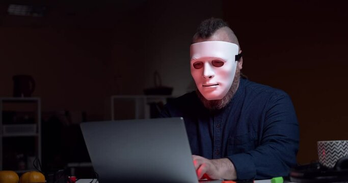 Bearded man in a white mask sits at a laptop. Computer hacker.