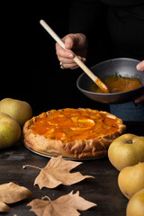 View of woman's hand with spoon in bowl with jam on apple pie on wooden table with apples and leaves, black background, vertical