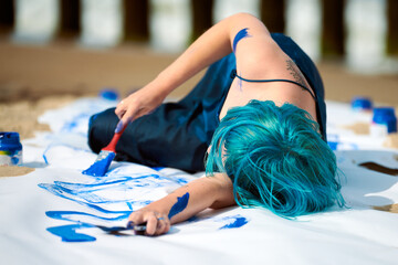 Artistic blue-haired woman performance artist smeared with gouache paints on large canvas on beach