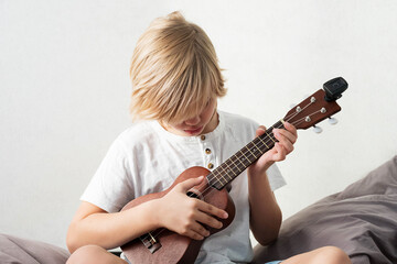 Young boy tuning ukulele at home. Blond haired boy sitting on couch playing acoustic guitar.