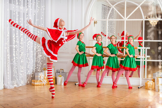 Children dressed as elves and practicing at a ballet barn look at a dancing ballerina dressed as santa claus in a spacious white studio.