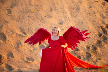 Fototapeta premium A plump blonde middle-aged woman with red angle wings in a desert with dunes and sand in a nice summer or spring sunny day with blue sky